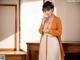 A woman in an orange kimono posing in front of a window.